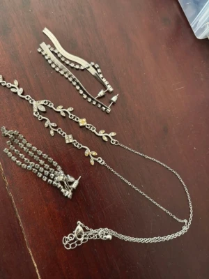 Smyckespaket med silverfärgade detaljer - Säljer ett smyckespaket med flera silverfärgade accessoarer. Innehåller halsband med bladformade detaljer och små stenar, armband med gnistrande stenar, örhängen och ett hängsmycke med kedjor. Materialet är metall med glittrande stenar i olika former.