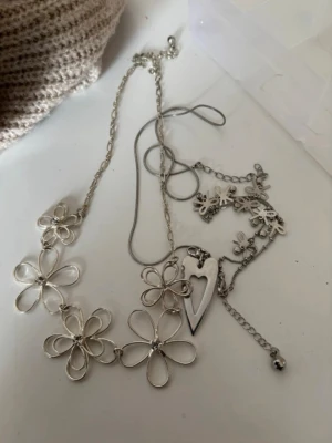 Silvriga halsband med blommor och hjärta - Två silvriga halsband, ett med stora blommor i metalltråd och ett med små blommor och ett hjärthänge. Båda har tunna kedjor och detaljerade blommotiv som ger en lekfull och trendig look. Ett armband med små rosetter som detalj. Perfekt för att addera en söt touch till din outfit.