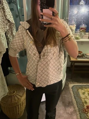 Rutig beige vindjacka med huva - Snygg beige vadderad vår/sommar jacka med rutigt mönster och guldfärgade knappar, fungerar även som kofta. Jackan har huva med snörning, dragkedja och knappar framtill samt korta ärmar. Perfekt för dig som vill ha en unik och trendig look. Skriv ifall ni vill ha mer bilder.