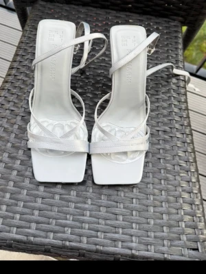 Vita sandaletter med remmar från Primark - Säljer ett par vita sandaletter från Primark med tunna, glansiga remmar och fyrkantig tå. Sandalerna har en stilren design med justerbart spänne runt ankeln och låg klack. Perfekta för dig som gillar minimalistisk och trendig stil. Använda en gång 