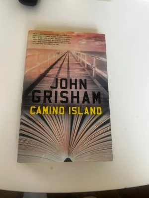 Camino Island av John Grisham - Spännande roman av John Grisham med omslag i varma toner och motiv av en brygga och en uppslagen bok. Boken är på svenska och handlar om stulna manuskript, en bokhandel och litterära mysterier på Camino Island i Florida.