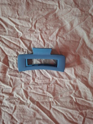 Blå hårklämma i matt plast - Säljer en stilren hårklämma i matt blå plast. Klämman har en rektangulär form med öppet mittparti och breda kanter, perfekt för att hålla upp håret på ett snyggt sätt. Enkel att matcha med olika outfits och superpraktisk till vardags.