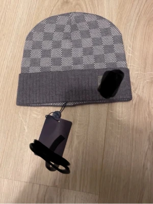 Grå mössa med rutmönster - Snygg grå mössa med diskret rutmönster och vikt kant. Mössan har en liten etikett på sidan och är tillverkad i mjukt stickat material som känns bekvämt på huvudet. Perfekt accessoar för kyliga dagar.