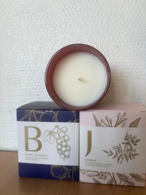 Set med doftljus i glas - Säljer ett set med tre doftljus i glasbehållare. Dofterna är Apple & Cinnamon i röd glasburk, Berry Blossom i blå förpackning och Jasmine i ljusrosa förpackning. Ljusen har dekorativa förpackningar med gulddetaljer och illustrationer av frukter och blommor.