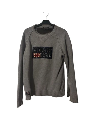 Grå tröja från Morris med tryck - Snygg grå tröja från Morris med broderat tryck på bröstet där det står 'Morris 1968' och en brittisk flagga. Tröjan har rund halsringning, ribbade muddar och är tillverkad i mjukt material. Perfekt för en avslappnad och stilren look.