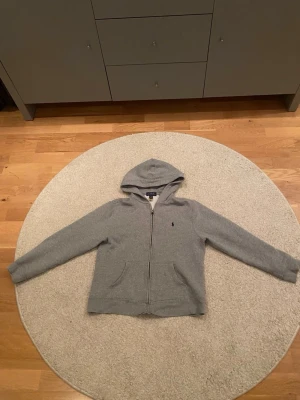 Grå hoodie från Polo Ralph Lauren - Klassisk grå hoodie från Polo Ralph Lauren med dragkedja framtill och huva. Tillverkad i mjuk bomullsmix och har den ikoniska lilla loggan broderad på bröstet. Perfekt för en chill och avslappnad stil.
