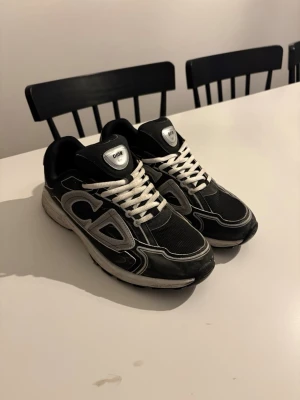 Svarta Dior B30 sneakers - Säljer ett par svarta Dior B30 sneakers med vita och grå detaljer. Skorna har en chunky sula, mesh och läderpaneler samt vita skosnören. Stora CD-loggor på sidorna och reflekterande detaljer ger en riktigt modern vibe.