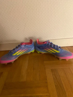 Adidas F50 blå/lila fotbollsskor - Säljer ett par Adidas F50 fotbollsskor i blå/lila med rosa och gula detaljer. Skorna har snörning, låg profil och yttersula med dobbar för bra grepp på planen. Tillverkade i syntetmaterial för lätt vikt och snabbhet. Perfekta för dig som vill sticka ut på fotbollsplanen.