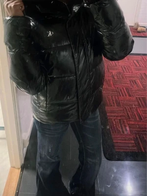 Svart pufferjacka från Calvin Klein - Säljer en svart, glansig pufferjacka från Calvin Klein med hög krage och dragkedja framtill. Jackan har en oversized look och CK-logga på bröstet. Perfekt för kalla dagar och riktigt snygg till jeans eller hoodies. Den är i en dam modell men passar även killar. Nypris ca 1200