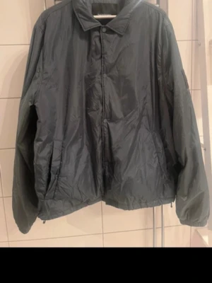 Svart vindjacka med patch - Snygg svart vindjacka med krage och dragkedja samt tryckknappar framtill. Jackan har två sidofickor och en diskret svart patch på ärmen. Tillverkad i ett glansigt, lätt material som står emot vind. Perfekt för dig som vill ha en stilren och enkel look.