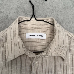 Randig skjorta från Samsøe Samsøe - Beige och vit randig skjorta från Samsøe Samsøe med klassisk krage och knäppning framtill. Skjortan har lång ärm och bröstficka, perfekt för en avslappnad men stilren look.