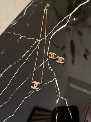 Chanel halsband  - Säljer ett elegant halsband från Chanel i guldton med den ikoniska CC-loggan dekorerad med små glittrande pärlor. Kedjan är tunn och smidig, perfekt för att lyfta vilken outfit som helst.