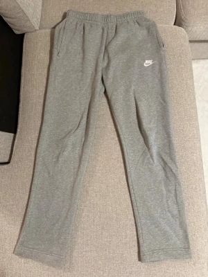 Gråa mjukisbyxor från Nike - Säljer ett par gråa mjukisbyxor från Nike med klassisk logga på vänster ben. Byxorna har resår i midjan, raka ben och sidofickor. Tillverkade i mjukt och bekvämt material, perfekta för chill dagar eller träning.