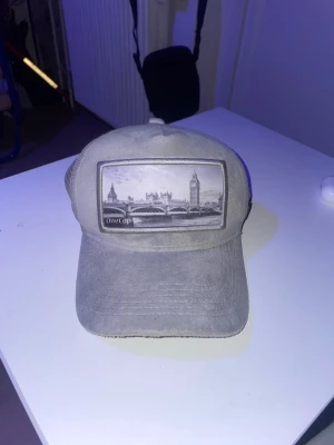 Grå velvet keps från onecap med Londonmotiv - Säljer en grå keps från Onecap med mesh på sidorna och ett stort tygmärke framtill som visar ett svartvitt motiv av London med Big Ben och Tower Bridge. Kepsen har böjd skärm och justerbar baksida för skön passform. Pris kan diskuteras.
