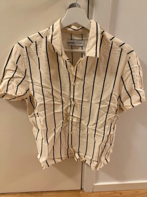 Randig kortärmad skjorta Bershka - Snygg kortärmad skjorta från Bershka i beige med svarta vertikala ränder. Skjortan har klassisk krage, knappar framtill och är tillverkad i ett lätt bomullsmaterial. Perfekt för en avslappnad och trendig look. Skjortan är aldrig använd och behöver strykas. 
