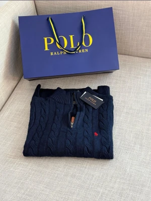 Mörkblå kabelstickad tröja Polo Ralph Lauren - Kabelstickad tröja i mörkblått från Polo Ralph Lauren med klassisk röd logobrodyr på bröstet. Tröjan har rund halsringning, långa ärmar och ribbade muddar. Har ej zip Tillverkad i mjukt material som ger en exklusiv känsla.