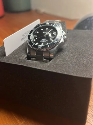 Svart och silver BiDen herrklocka - Stilren herrklocka från BiDen med svart urtavla och silverfärgat armband i metall. Klockan har tydliga vita markeringar, datumvisning och vridbar bezel. Quartz-urverk och vattentät ner till 30 meter. Perfekt accessoar för dig som gillar klassisk design.