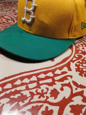 Gul och grön keps med B-logga - Snygg keps i gult med grön skärm och en vit broderad B-logga framtill. Klassisk basebollmodell med böjd skärm och justerbar passform. Perfekt accessoar för en sportig och avslappnad stil.