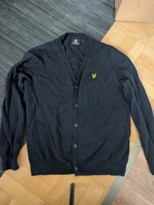 Svart cardigan från Lyle & Scott stl L - Klassisk svart cardigan från Lyle & Scott med gul broderad logga på bröstet. Tröjan har v-ringning, knäppning framtill med fem knappar och ribbade muddar. Perfekt för lager på lager och enkel att matcha med olika outfits.