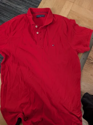 Röd pikétröja från Tommy Hilfiger stl M - Snygg röd pikétröja från Tommy Hilfiger med klassisk krage och två knappar framtill. Custom fit-modell med diskret logga på bröstet. Perfekt till jeans eller shorts för en clean och stilren look.
