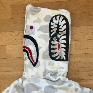 A BATHING APE camo shark-hoodie med dragkedja - Säljer en BAPE zip hoodie i vit och grå camo med hajmotiv på huvan. Hoodien har dragkedja hela vägen upp, två fickor fram och detaljerade broderier på huvan. Ikonisk streetwear-stil och coolt camomönster. Perfekt för dig som vill sticka ut.