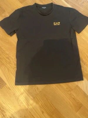 Svart EA7 t-shirt med guld logga - Snygg svart t-shirt från EA7 med gul logga på bröstet och gula ränder på ryggen. Klassisk passform och rund halsringning. Perfekt till jeans eller shorts för en clean och stilren look.