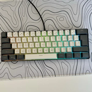 RGB mekaniskt tangentbord 60% - Säljer ett kompakt mekaniskt tangentbord med RGB-belysning och 60% layout. Tangentbordet har vita och gråa tangenter med tydliga symboler och är anslutet via kabel. Perfekt för gaming eller produktivitet, och har en modern design med plastmaterial.