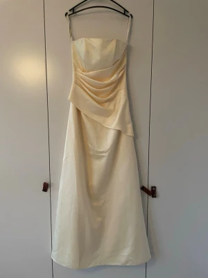 Lång ljusgul balklänning - Elegant långklänning perfekt för balen i en ljusgul färg och draperad front. Klänningen har subtila pärldetaljer upptill och är tillverkad i ett glansigt material som påminner om satin. Dragkedja i ryggen för enkel påklädning. Perfekt för speciella tillfällen. Aldrig använd. Köpt för 1900kr.