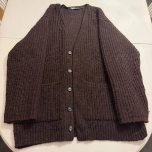 Brun stickad kofta från Our Legacy - Brun stickad kofta med v-ringning och fem knappar framtill från Our Legacy. Koftan har två stora fickor på framsidan och en avslappnad passform. Perfekt för lager på lager och enkel att matcha med olika outfits. 