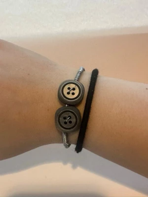 Silverarmband med gråa knappar - Armbandet är gjort av silverfärgad metalltråd med en mindre metalltråd virad runt. Fäst till metalltråden är det två gråa knappar. Armbandet är dessutom justerbart så att du kan anpassa det till din storlek.