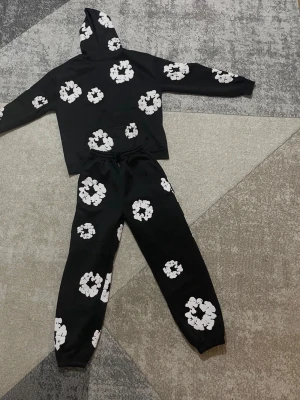 Denim Tears Hoodie & Pants - Svart hoodie med stora vita blommönster över hela plagget. Hoodien har en klassisk huva och en magficka framtill. Tillverkad i mjukt material som passar perfekt för en avslappnad och trendig look.