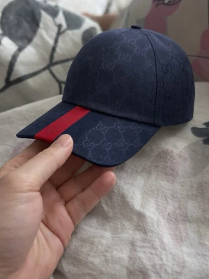 Mörkblå Gucci keps med röd rand - Snygg mörkblå keps från Gucci med diskret GG-monogram över hela tyget och en bred röd rand på skärmen. Klassisk böjd skärm och justerbar passform. Tillverkad i slitstarkt tyg som ger en exklusiv känsla.