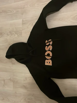 Svart hoodie från BOSS med tryck - Snygg svart hoodie från BOSS med stort beige och vitt BOSS-tryck på bröstet. Klassisk huva med dragsko och lång ärm. Perfekt för en avslappnad och trendig stil. Materialet ser ut att vara mjukt och bekvämt, troligen bomull eller bomullsblandning.