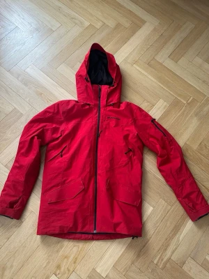 Röd skidjacka Peak Performance - Snygg röd skidjacka från Peak Performance med huva och svarta detaljer. Jackan har dragkedja framtill, flera fickor med dragkedja och justerbar huva. Materialet är slitstarkt och vattenavvisande, perfekt för blåsiga dagar och i skidbacken 