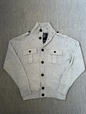 Ljusgrå stickad cardigan med fickor - Snygg ljusgrå stickad cardigan från Morris med svarta knappar, två bröstfickor med lock och knappar, samt ribbade muddar och nederkant. Axelpartiet har dekorativa axelklaffar och ärmarna har förstärkta armbågslappar. Perfekt till lager på lager-stil.