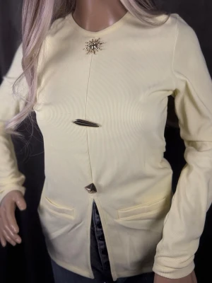 Ljusgul ribbad kofta med dekorativa knappar - Ljusgul ribbad kofta med lång ärm och två framfickor. Koftan har unika dekorativa knappar i metall och en snygg broschdetalj vid halsen. Perfekt för dig som gillar stilrena plagg med en twist.