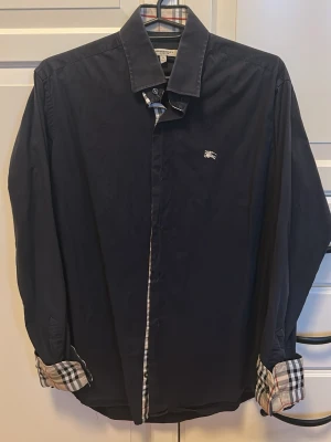 Mörkblå skjorta från Burberry - Snygg mörkblå skjorta från Burberry med klassiskt rutigt tyg på insidan av manschetterna och knappslån. Skjortan har lång ärm, krage och en liten broderad Burberry-logga på bröstet. Perfekt för dig som gillar stilrena detaljer och ikonisk design.