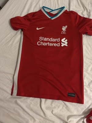 Liverpool matchtröja Nike röd - Säljer en röd Liverpool FC matchtröja från Nike med vitt Standard Chartered-tryck på bröstet och spelarnamn Elliott samt nummer 67 på ryggen. Tröjan har turkosa och vita detaljer vid kragen och är tillverkad i ett lätt, andningsbart material. Storlek 147-158.