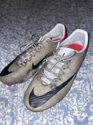Nike Mercurial fotbollsskor vit/röd - Säljer ett par Nike Mercurial fotbollsskor i vitt med svarta detaljer och röd sula. Skorna har snörning, låg profil och är tillverkade i syntetmaterial för bra grepp och kontroll på planen. Perfekta för dig som vill ha lätta och snabba skor till match eller träning.