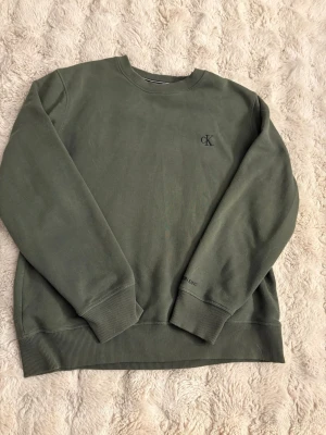 Olivgrön sweatshirt från Calvin Klein - Snygg olivgrön sweatshirt från Calvin Klein Jeans med diskret CK-logga broderad på bröstet. Tröjan har rund halsringning, ribbade muddar vid ärmslut och nederkant samt mjuk insida. Perfekt för en avslappnad och stilren look.