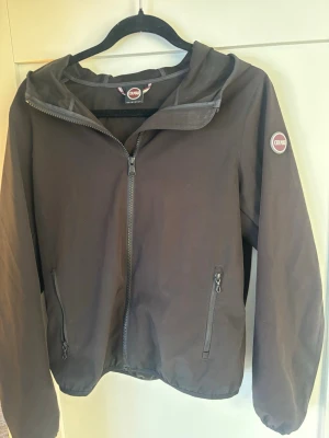 Svart vindjacka från Colmar - Snygg svart vindjacka från Colmar Originals med huva och hel dragkedja framtill. Jackan har två fickor med dragkedja och Colmar-logga på ärmen. Tillverkad i ett lätt och vindtätt material, perfekt för blåsiga dagar.