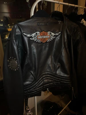 Harley-Davidson svart skinnjacka - Snygg svart skinnjacka från Harley-Davidson med broderad logga och vingar på ryggen. Klassisk bikerstil med dragkedjor, bälte i midjan och detaljerade sömmar. Perfekt för dig som vill ha en tuff och ikonisk look.