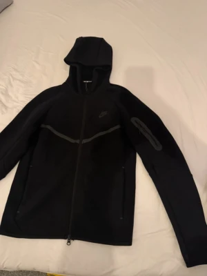 Svart Nike Tech Fleece hoodie - Säljer en svart Nike Tech Fleece hoodie med dragkedja och huva. Hoodien har diskret Nike-logga på bröstet, fickor med dragkedja och en extra ficka på ärmen. Tillverkad i mjukt och flexibelt material som är perfekt för chill eller träning.