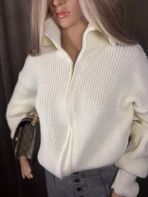 Vit stickad ziptröja med krage S-M - Säljer en stilren vit stickad tröja med dragkedja och hög krage. Tröjan har en chunky ribbstickad struktur och långa ärmar med breda muddar. Perfekt till jeans för en clean look. Materialet känns mjukt och värmande.