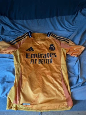 Gul Real Madrid matchtröja Adidas - Snygg gul Real Madrid matchtröja från Adidas med mörkblå detaljer och klubbmärke på bröstet. Tröjan har trycket 'Emirates Fly Better' på framsidan och klassiska Adidas-ränder på axlarna. Perfekt för fotbollsträning eller att bära till vardags.