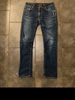 Mörkblå jeans från Nudie Jeans - Säljer ett par mörkblå jeans från Nudie Jeans med klassisk femficksmodell och snygga slitningar. Jeansen har kontrastsömmar, raka ben och detaljerade bakfickor med Nudie-loggan. Tillverkade i denim av bomull.