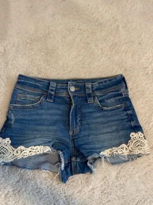 Blå jeansshorts med spetsdetaljer - Snygga blå jeansshorts med råa kanter och vita spetsdetaljer längs bensluten. Klassisk femficksmodell med bälteshällor och knappstängning. Perfekta för sommardagar och ger en cool, avslappnad vibe. Storlek 152.