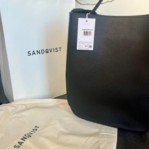 Svart väska från Sandqvist - Hej! Säljer en riktigt schysst helt ny väska från Sandqvist Bags. Fick denna i utbyte mot ett modell jobb, lappen är kvar på väskan, påsen ingår. Funkar som unisex! Nypris 4495kr, information finns nedan.  Höjd: 38.5 cm Bredd: 32 cm Djup: 12 cm Kapacitet: 14.7 L  Denna hobo tote har en innerficka med dragkedja och två pennfickor. Ett magnetlås för huvudfacket säkerställer snabb och säker åtkomst till dina saker.   Det rymliga huvudfacket har en kapacitet på 14,7 liter och rymmer laptops upp till