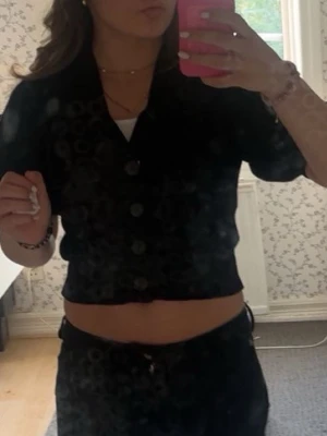 Svart kortärmad cardigan crop top - Trendig svart cardigan crop top med korta ärmar och stora knappar framtill. Modellen är croppad och har en klassisk krage. Perfekt att styla med höga byxor eller kjol för en cool look.