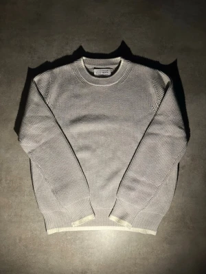 Grå stickad tröja Maison Margiela - Snygg grå stickad tröja från Maison Margiela med rund halsringning och ribbade muddar. Tröjan har en klassisk rak passform och är tillverkad i ett mjukt, strukturerat material som känns lyxigt och stilrent.